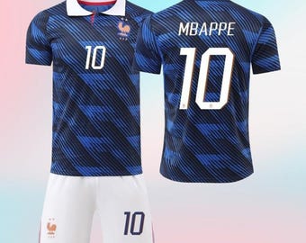 May include: A dark blue football shirt with a white collar, featuring a diagonal striped pattern, the number 10, and a rooster emblem. The back of the shirt displays the name MBAPPE and the number 10. White football shorts with the number 10 are included.