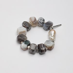 May include: A bracelet featuring faceted, banded agate stones in shades of gray, brown, and white, interspersed with small silver-toned spacers. A single irregular, cream-colored pearl is attached near a silver tag engraved with the text PURAPERIA.
