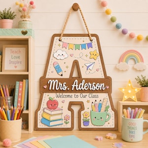 May include: A wooden classroom door sign shaped like the letter A, featuring the text Mrs. Aderson and Welcome to Our Class. The sign is decorated with colorful illustrations of a smiling apple on books, a pencil, a paper plane, and a mug with pens.