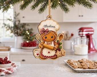 May include: A flat wooden Christmas ornament shaped like a gingerbread person wearing a chef hat and apron, holding a rolling pin, whisk, and wooden spoon. The name Madison is printed on the hat. It features red and white accents and a jute string.