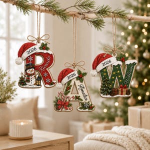 May include: Three hanging Christmas ornaments shaped as the letters R, A, and W. Each letter features a red Santa hat, the year 2026, and a name: Ricardo, Amelia, and William. The ornaments are decorated with snow, holly, and festive motifs.