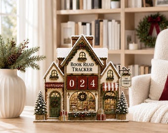 May include: A decorative tabletop book read tracker shaped like a miniature shop with a snowy roof, featuring the text "BOOK READ TRACKER" above three red number tiles displaying "0 2 4" and a hanging sign that reads "THE ANDERSON FAMILY".