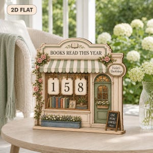 May include: A decorative wooden book tracker shaped like a storefront with the text BOOKS READ THIS YEAR and Violet's Bookshop. It features a green striped awning, a green door, pink roses, a blue flower box, and three hanging number tags displaying 158.