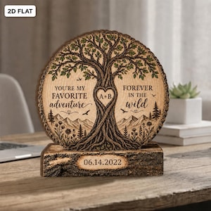 May include: A circular wooden plaque featuring a laser-engraved tree with green leaves, mountains, and birds. The text reads: YOU'RE MY FAVORITE ADVENTURE FOREVER IN THE WILD, with A+B inside a heart. The base displays the date 14.06.2022.