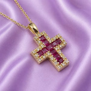 Peut inclure: Un pendentif croix en or monté sur une chaîne fine, orné de rubis rouges de taille carrée au centre, entourés d'une bordure de petits diamants blancs ronds et brillants. L'ensemble présente une finition métallique polie et éclatante.