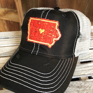 May include: A black and gray trucker hat with a red and gold state of Iowa patch with a gold heart in the center. The hat has a distressed look with frayed edges.