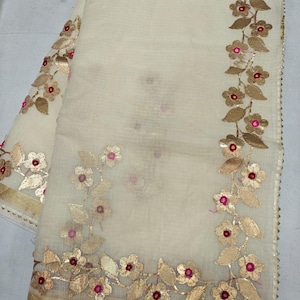 May include: A cream-colored sheer fabric textile featuring an embroidered floral vine pattern. The design includes metallic gold leaves and flowers with small, vibrant pink circular centers. A solid gold border runs along the bottom edge.