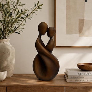 May include: A dark brown wooden sculpture depicting two abstract figures in an embrace, featuring smooth, flowing curves and rounded heads. The sculpture has a solid, continuous form that tapers from a wide, circular base to the two heads at the top.