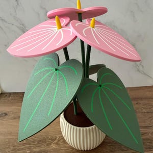 May include: A decorative artificial plant featuring three pink heart-shaped anthurium flowers with yellow spadices and white line details. The plant has several large, textured green leaves with light green veins, all set in a white ribbed pot.