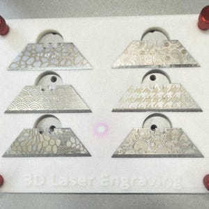 May include: A white rectangular base with four red corner knobs holds six trapezoidal metal utility blades. Each blade features a distinct laser-engraved pattern on its surface. The base is embossed with the text 3D Laser Engraving.