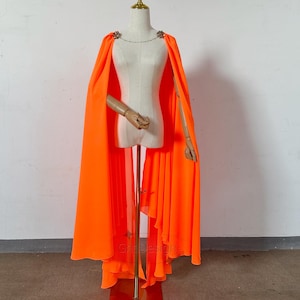 May include: A bright neon orange floor-length cape made of flowing, lightweight fabric. The cape features a delicate gold-tone chain closure at the neckline, adorned with two small, circular, multi-colored jeweled embellishments on each side.