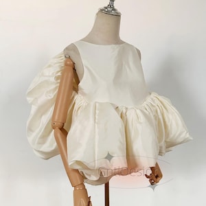 May include: A sleeveless cream-colored top featuring a round neckline and voluminous, gathered puff sleeves. The garment is constructed from a smooth, slightly lustrous fabric and is displayed on a mannequin. The text GntlDesign is visible.