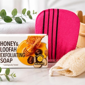 Könnte beinhalten: Eine rechteckige Schachtel mit Honig- und Luffa-Peelingseife mit einem Nettogewicht von 100g, verziert mit Honig und Kastanien. Daneben liegen ein leuchtend pinker Peelinghandschuh mit drei schwarzen Streifen und ein beiger Luffaschwamm.