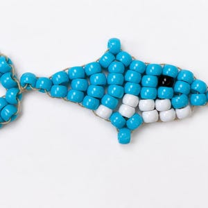 May include: A dolphin-shaped ornament crafted from bright blue plastic pony beads strung together on a thin cord. The design features a single black bead for the eye and a row of white beads along the underside to represent the belly area.