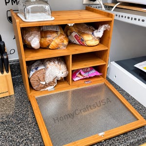 May include: A multi-level bamboo bread box with a drop-down door featuring a ribbed, translucent panel. The interior contains multiple shelves holding various packaged baked goods, with a clear glass butter dish on the top surface.