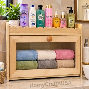May include: A light-colored wooden storage cabinet with a clear front panel and a round knob. Inside, six folded towels are stacked in blue, white, pink, green, gray, and tan. The top shelf holds various shampoo and oil bottles.