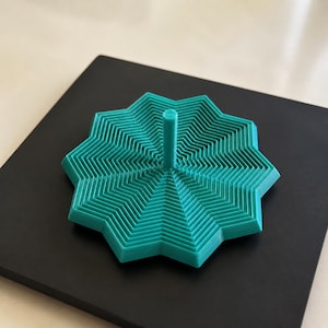 May include: A teal, star-shaped object with a multi-layered, concentric, zigzagging pattern radiating from a central vertical cylinder. The object features a series of stepped, geometric ridges that create a textured, three-dimensional appearance.