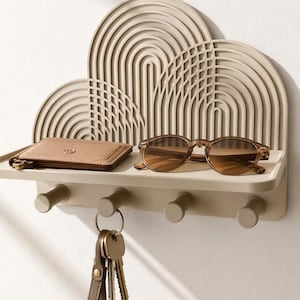 May include: A beige wall-mounted shelf featuring a decorative back panel with concentric arch patterns. The shelf includes four cylindrical hooks underneath, currently holding a set of keys on a leather fob, with a wallet and sunglasses resting on top.