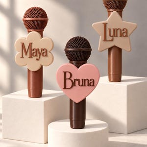 May include: Three brown microphones featuring custom name attachments. One has a beige flower shape with the name Maya, one has a pink heart shape with the name Bruna, and one has a beige star shape with the name Luna in brown lettering.