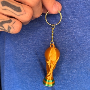 May include: A gold-colored trophy keychain featuring a spherical top and a tapered base with a green band. The trophy is attached to a gold-toned metal split ring by a short chain. The object has a smooth, metallic finish and a distinct, molded shape.