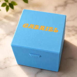 May include: A bright blue square box with a smooth matte finish. The top surface features the name GABRIEL embossed in a bold, yellow, rounded font. The box has a small rectangular latch on the front side for opening and closing the lid.