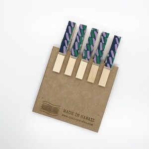 May include: Set of six wooden clothes pegs with fabric covers. The fabric is a geometric pattern in shades of purple, green, and blue. The clothes pegs are arranged on a brown card with the text "MADE IN HAWAII" and a website address.