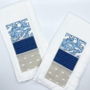 May include: Two white washcloths with a patchwork design featuring blue and beige fabric. The fabric has a pattern of sharks and geometric shapes.