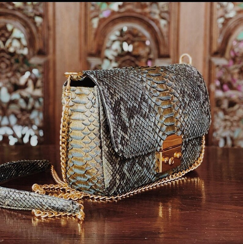 K&ouml;nnte beinhalten: Eine strukturierte Umh&auml;ngetasche mit einem dunkelgrauen und goldfarbenen Schlangenledermuster. Die Tasche verf&uuml;gt &uuml;ber einen goldfarbenen Metallkettenriemen und einen rechteckigen goldfarbenen Metallverschluss an der Frontklappe sowie einen passenden Riemen.