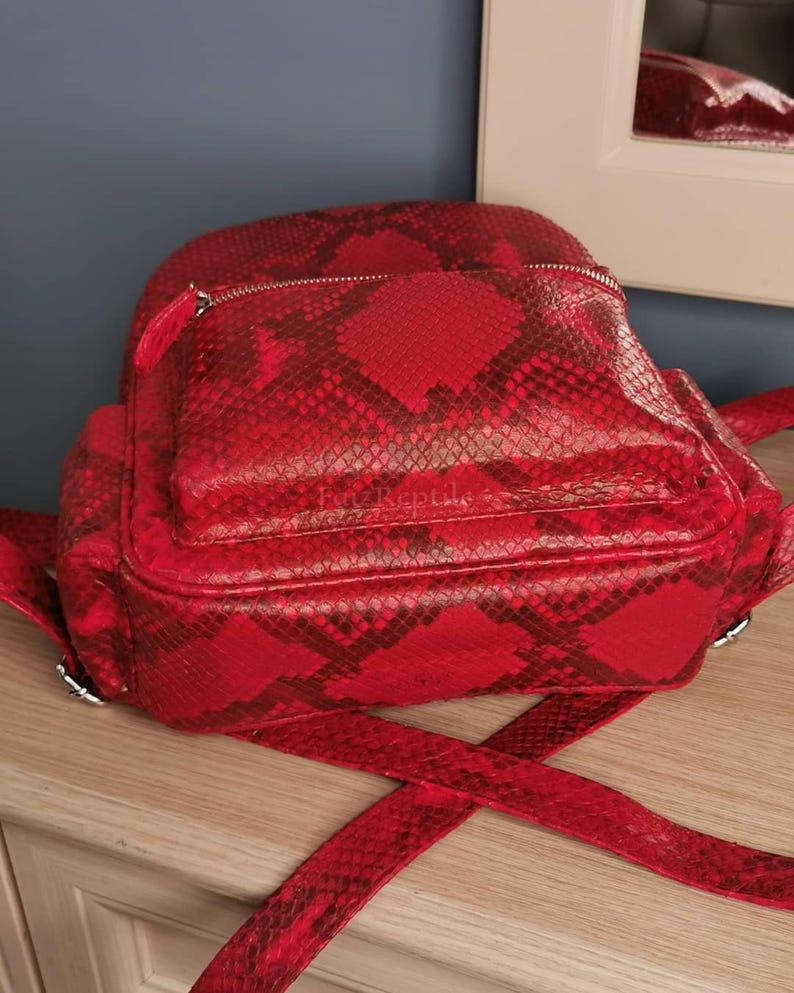 May include: A bright red backpack featuring a textured snakeskin pattern throughout. The bag includes a front zippered pocket with a silver-toned metal zipper and the text FanzReptile embossed on the front panel. It has adjustable shoulder straps.