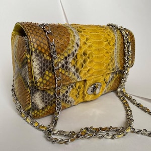 May include: A rectangular shoulder bag featuring a textured snakeskin pattern in vibrant yellow, gray, and white tones. The bag includes a silver-toned metal chain strap and a matching silver-toned twist-lock closure on the front flap.