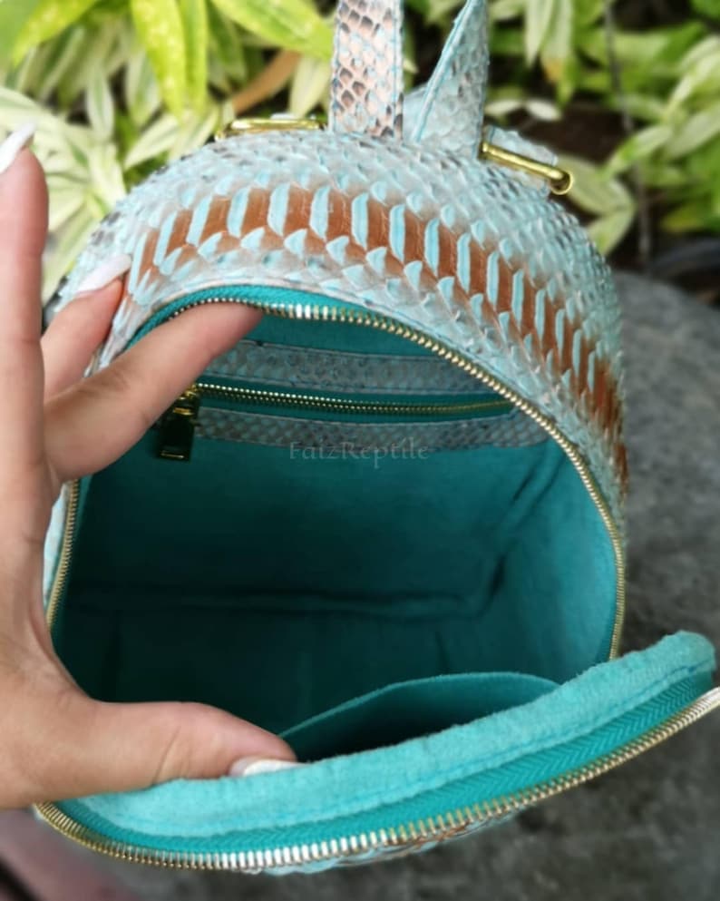 May include: A small backpack with a textured, light blue and brown snakeskin-patterned exterior. The interior is lined with solid teal fabric and features a zippered pocket. The word FatzReptile is printed in small, light text inside the bag.
