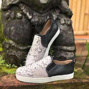 May include: A pair of slip-on trainers featuring a textured snakeskin pattern in shades of grey and white on the vamp, contrasted with solid black leather side panels, black elastic gussets, and a thick white rubber sole for a clean finish.