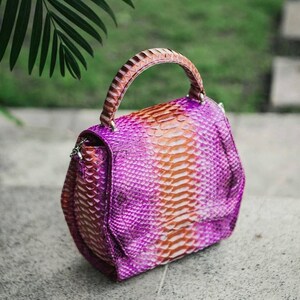 Puede incluir: Un bolso estructurado con un patrón de piel de serpiente texturizado en un degradado vibrante de tonos morados y naranjas. El bolso incluye un asa superior curva y un cierre de solapa, mostrando una textura de escamas en toda la superficie.