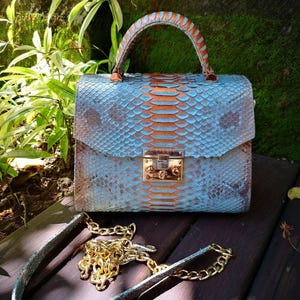 May include: A structured handbag featuring a textured snakeskin pattern in shades of light blue and copper. The bag includes a top handle, a metallic silver-tone clasp closure, and a detachable gold-tone chain strap with matching snakeskin accents.