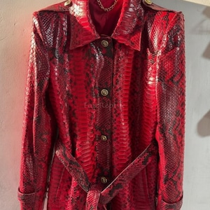 May include: A long-sleeved red and black snakeskin-patterned trench coat featuring a pointed collar, shoulder epaulets, a matching tie belt, and gold-toned buttons down the front. The interior lining is red and includes a gold-toned chain hanger.