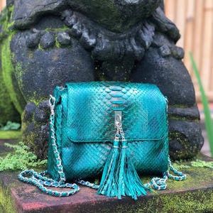 Puede incluir: Un bolso de mano con textura de piel de serpiente en color verde azulado vibrante, con cierre de solapa rectangular, correa de cadena metálica plateada y un adorno de doble borla que cuelga de un cierre central plateado.