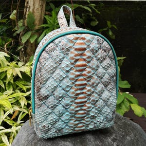 May include: A small backpack featuring a textured, scale-like pattern in shades of light blue, brown, and grey. The bag has a rounded top, a top handle, and a bright teal zip closure that runs along the perimeter of the front panel.