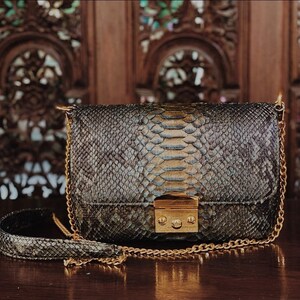 Peut inclure: Un sac à bandoulière rectangulaire présentant un motif en peau de serpent gris foncé et noir. Le sac comprend une fermeture à fermoir en métal doré sur le rabat avant et une bandoulière en chaîne dorée assortie avec une épaulette en cuir.