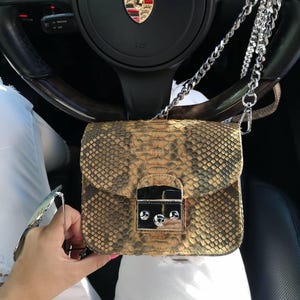 Puede incluir: Un bolso de hombro rectangular con un patrón de piel de serpiente texturizado en tonos bronceado, marrón y negro. El bolso tiene un cierre de metal plateado en la solapa frontal y una correa de cadena plateada sujeta a los lados con clips.