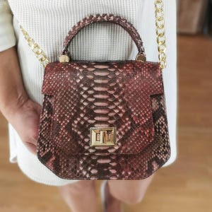 May include: A structured handbag featuring a textured snakeskin pattern in shades of deep red, brown, and black. The bag includes a top handle, a gold-toned metal turn-lock closure on the front flap, and a matching gold-toned metal chain shoulder strap.