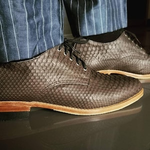 Può includere: Un paio di scarpe stringate grigio scuro con un motivo testurizzato simile alla pelle di serpente sulla tomaia, lacci neri e una suola color legno marrone chiaro con base del tacco nera, indossate con pantaloni gessati blu.