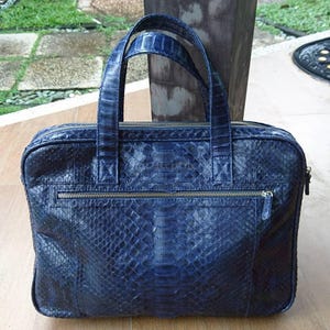 May include: A rectangular navy blue briefcase featuring a textured snakeskin pattern. The bag includes two top handles, a horizontal zippered front pocket, and a main zippered compartment with silver-toned metal hardware throughout the exterior.