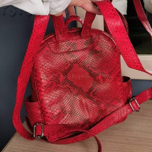 May include: A bright red backpack featuring a textured snakeskin pattern and a top handle. The bag includes adjustable shoulder straps with silver-toned metal buckles and side pockets. The text "FatzReptile" is visible on the front surface.