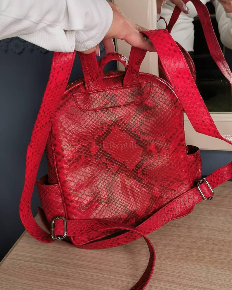 May include: A bright red backpack featuring a textured snakeskin pattern and a top handle. The bag includes adjustable shoulder straps with silver-toned metal buckles and side pockets. The text "FatzReptile" is visible on the front surface.