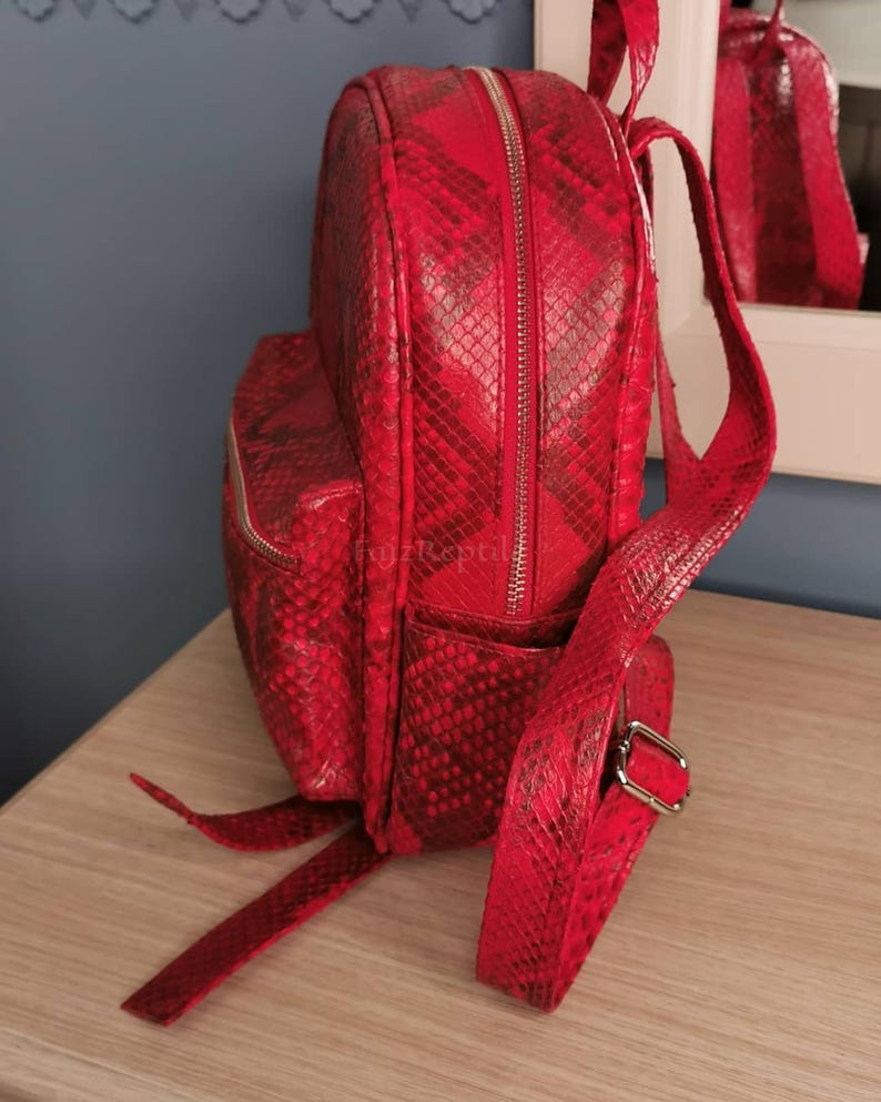 May include: A vibrant red backpack featuring a textured snakeskin pattern throughout. The bag includes a main zippered compartment, a smaller front zippered pocket, and adjustable shoulder straps with silver-toned metal hardware buckles.