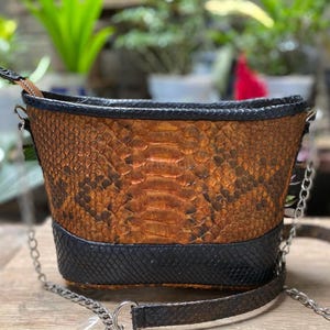 Peut inclure: Un sac à main de style seau présentant un motif en peau de serpent marron texturé sur le corps principal et une base en peau de serpent noire. Le sac comprend une bordure noire sur le bord supérieur et une bandoulière en chaîne métallique argentée.