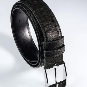 Op de afbeelding: Een zwarte riem met een gestructureerd, schubachtig patroon op het buitenoppervlak. De riem is voorzien van een gepolijste zilverkleurige metalen gesp en een bijpassende lus. De binnenzijde is gevoerd met glad zwart materiaal en heeft gaatjes.