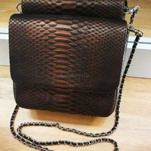 May include: A rectangular shoulder bag with a dark brown and black snakeskin pattern. The bag features a flap closure and a silver-toned metal chain strap interwoven with dark material. The text "FarReptile" is visible on the front flap.