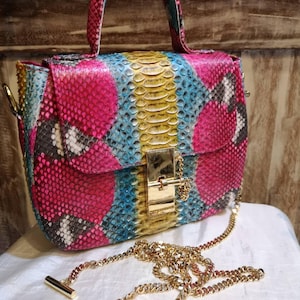 Peut inclure: Un sac à main structuré présentant un motif en peau de serpent aux couleurs vives, notamment rose vif, bleu éclatant et jaune. Le sac est doté d'une poignée supérieure, d'un fermoir métallique rectangulaire doré et d'une bandoulière en chaîne dorée.