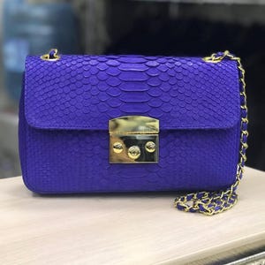 Op de afbeelding: Een levendige paarse rechthoekige schoudertas met een gestructureerd slangenleerpatroon. De tas is voorzien van een gepolijste goudkleurige metalen sluiting op de voorflap en een bijpassende goudkleurige kettingriem verweven met paars materiaal.